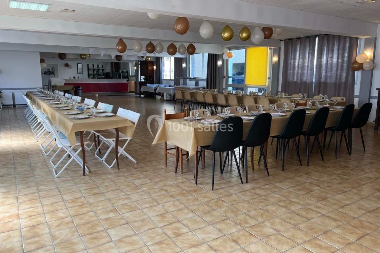 Salle de réception avec tables alignées, nappes beige, chaises variées et décorations suspendues en ballons.