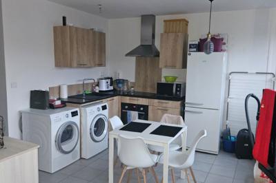 Cuisine moderne avec meubles en bois clair, électroménagers, table blanche et chaises, lave-linge et sèche-linge visibles.