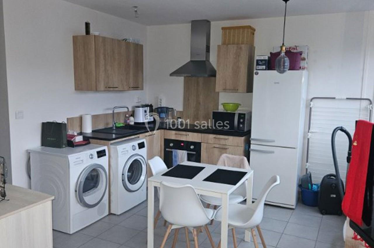 Cuisine moderne avec meubles en bois clair, électroménagers, table blanche et chaises, lave-linge et sèche-linge visibles.