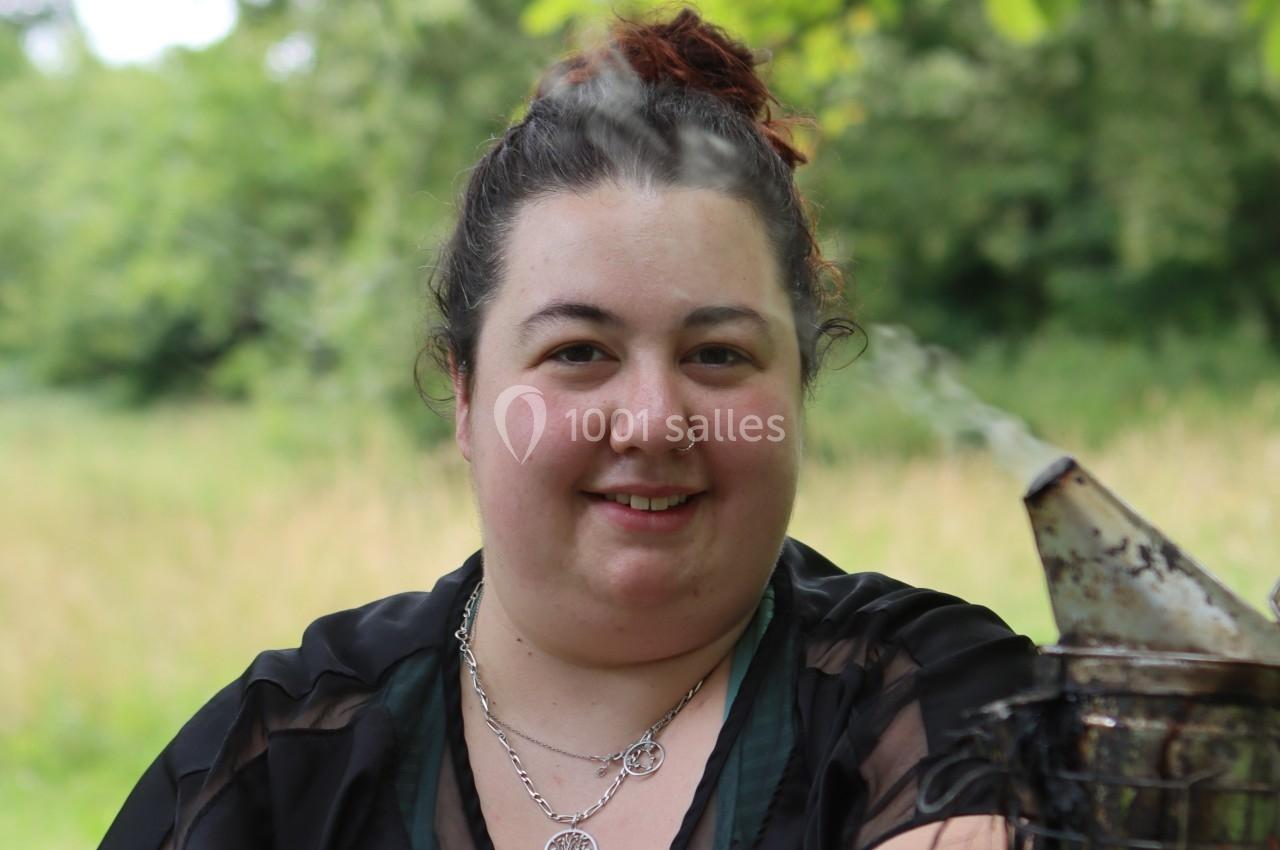 Femme souriante en extérieur, devant un fond de verdure, avec un enfumoir métallique visible au premier plan.