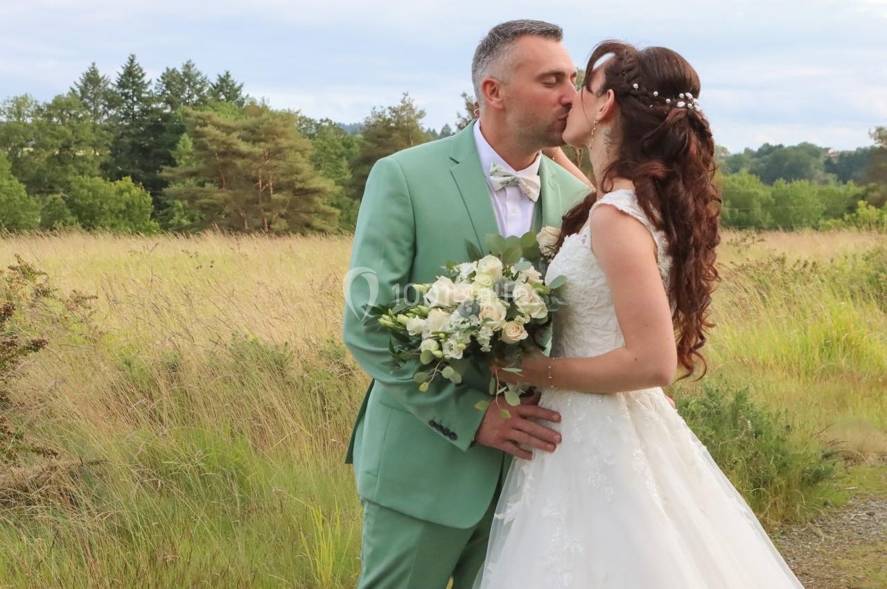 Un couple en tenue de mariage s'embrasse dans un champ verdoyant, le marié tenant un bouquet de fleurs blanches.