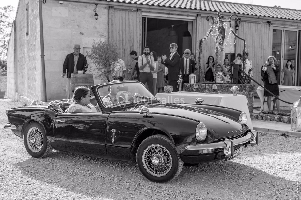 Une voiture décapotable ancienne noire arrive devant un bâtiment en pierre, des invités applaudissent en arrière-plan.