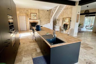 Façade d'une maison en pierre avec terrasse ensoleillée, chaises longues et pots de fleurs sur un sol pavé clair.