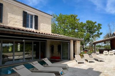 Façade d'une maison en pierre avec terrasse ensoleillée, chaises longues et pots de fleurs sur un sol pavé clair.