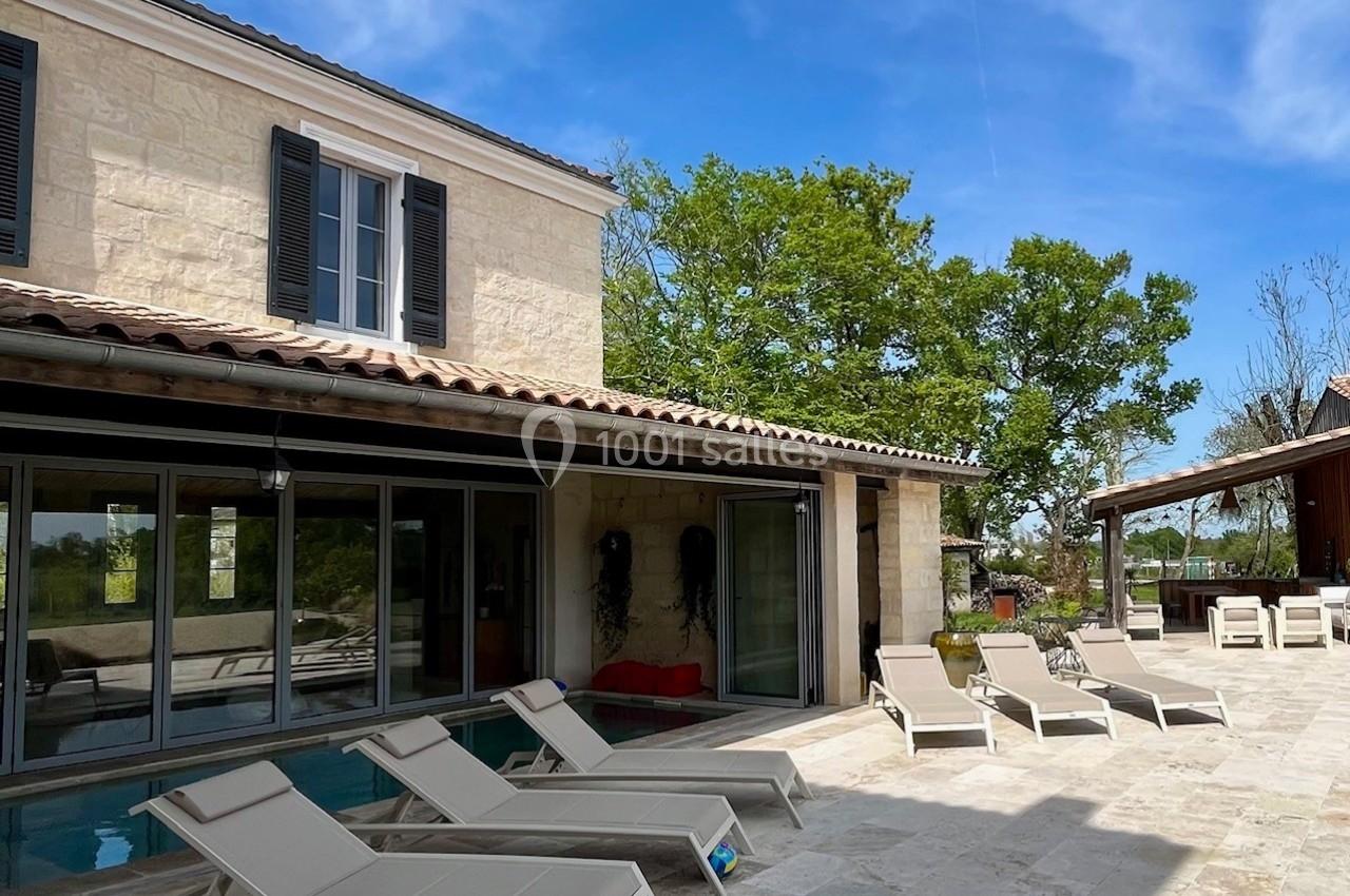 Maison en pierre avec terrasse, piscine intérieure visible à travers des baies vitrées, et transats sous un ciel bleu.
