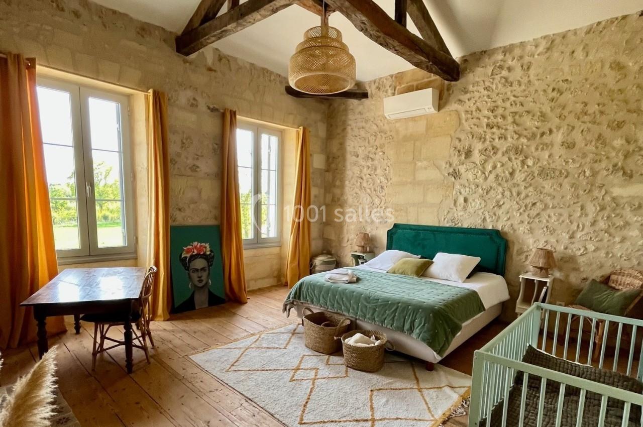 Chambre lumineuse avec murs en pierre, lit double, lit bébé, poutres apparentes, tapis et grandes fenêtres donnant sur l…