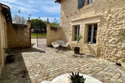 Façade d'une maison en pierre avec terrasse ensoleillée, chaises longues et pots de fleurs sur un sol pavé clair.