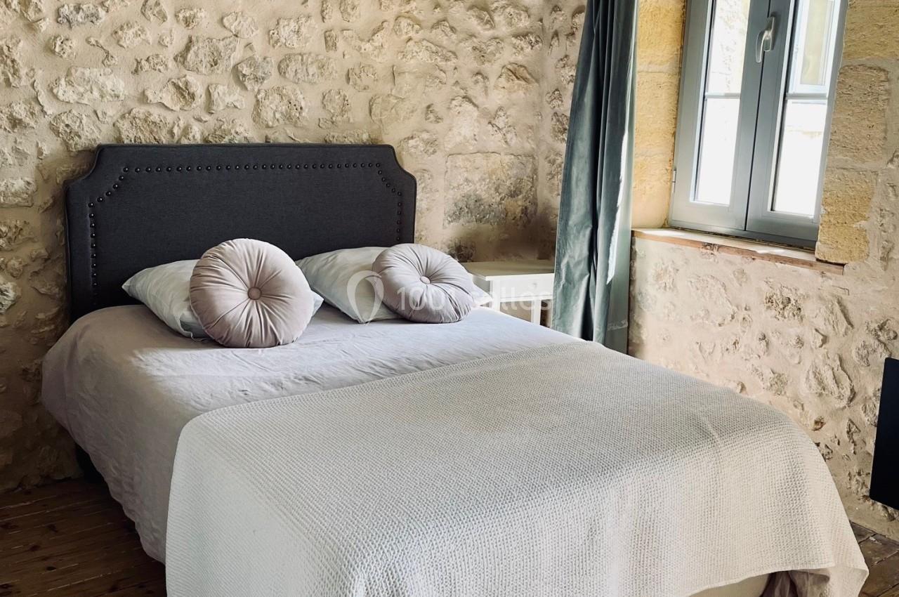 Chambre avec murs en pierre, lit double avec coussins décoratifs, parquet en bois et fenêtre avec rideaux verts.