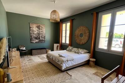 Façade d'une maison en pierre avec terrasse ensoleillée, chaises longues et pots de fleurs sur un sol pavé clair.