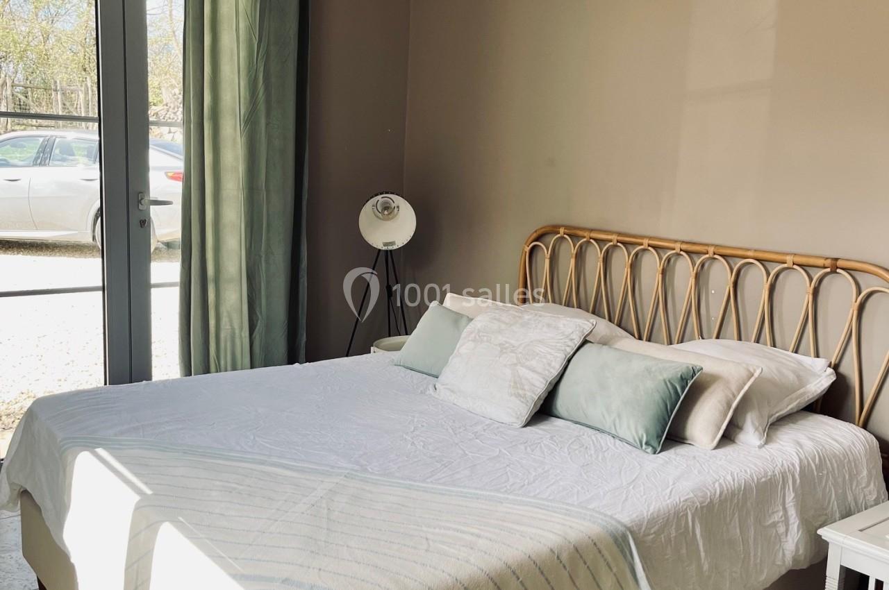 Lit double avec tête de lit en rotin, draps blancs et coussins pastel, dans une chambre lumineuse avec rideaux verts.