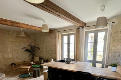 Façade d'une maison en pierre avec terrasse ensoleillée, chaises longues et pots de fleurs sur un sol pavé clair.