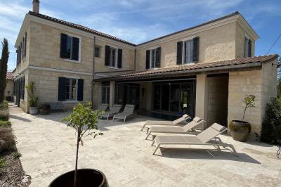 Façade d'une maison en pierre avec terrasse ensoleillée, chaises longues et pots de fleurs sur un sol pavé clair.