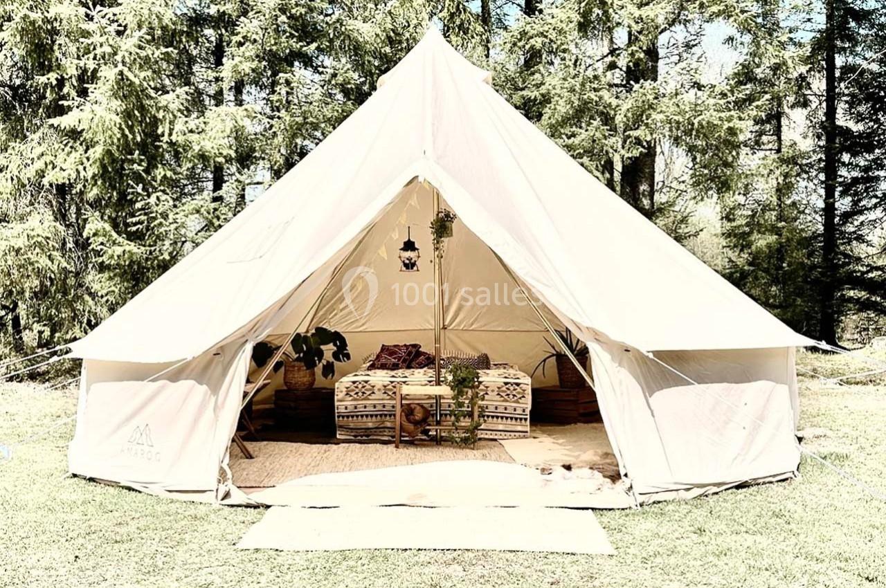 Tente beige en toile installée dans une clairière, aménagée avec des coussins et des plantes pour un espace cosy.