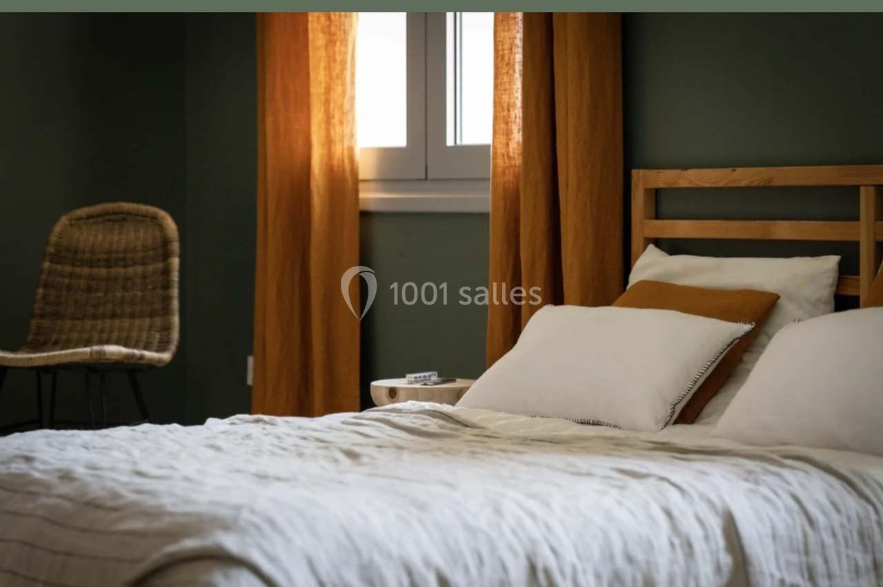 Lit avec draps blancs, coussins et tête de lit en bois, près d'une fenêtre encadrée de rideaux orange, chaise en rotin à…