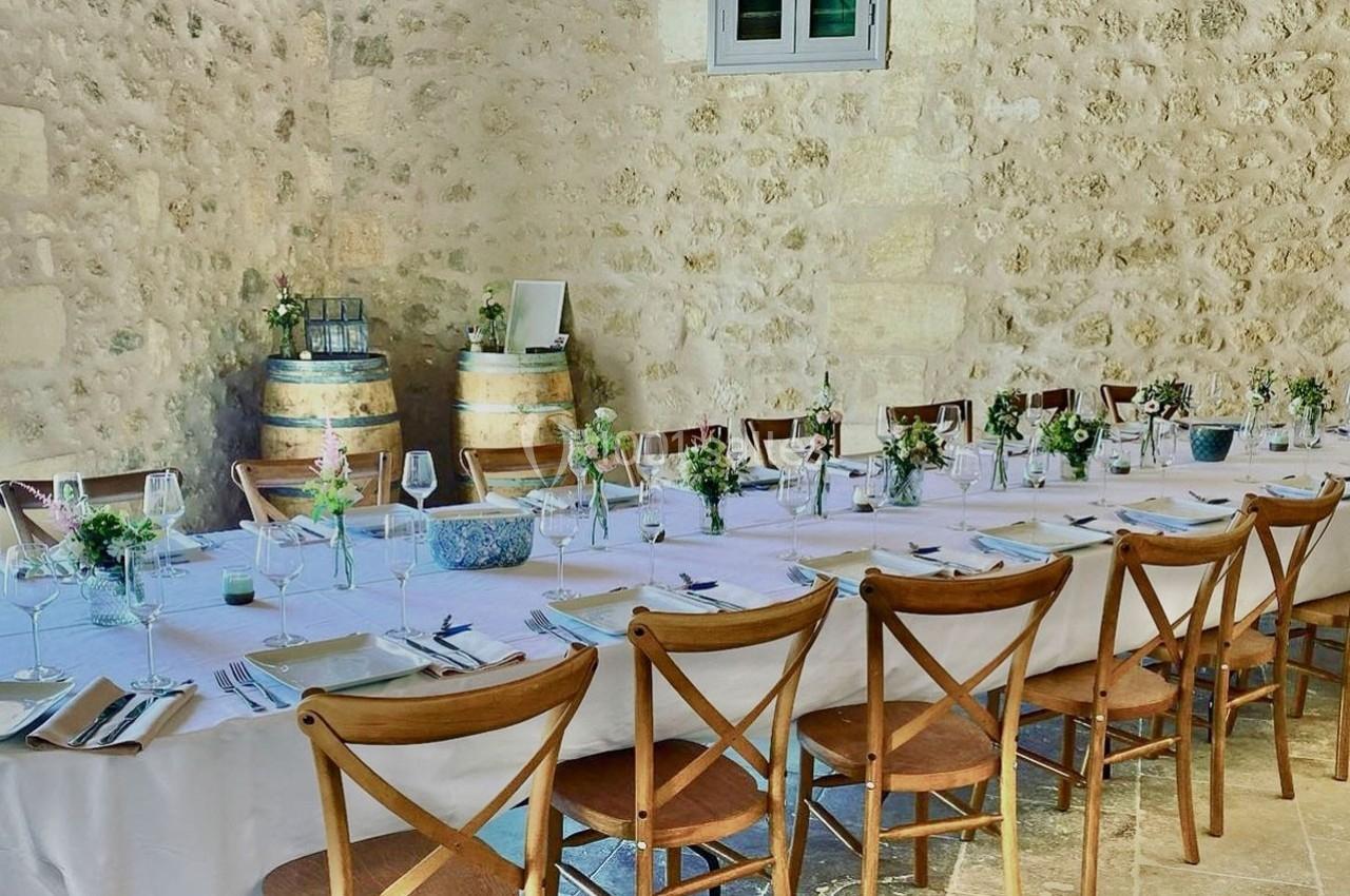 Grande table dressée avec nappes blanches, chaises en bois, vaisselle élégante et décorations florales dans une salle en…