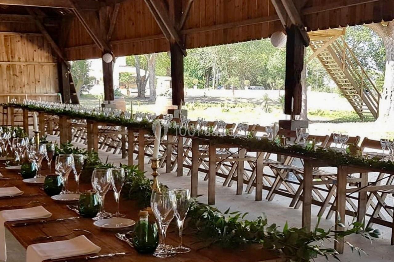 Grande table en bois décorée de verres, bougies et feuillage, installée dans une grange ouverte avec vue sur l'extérieur.
