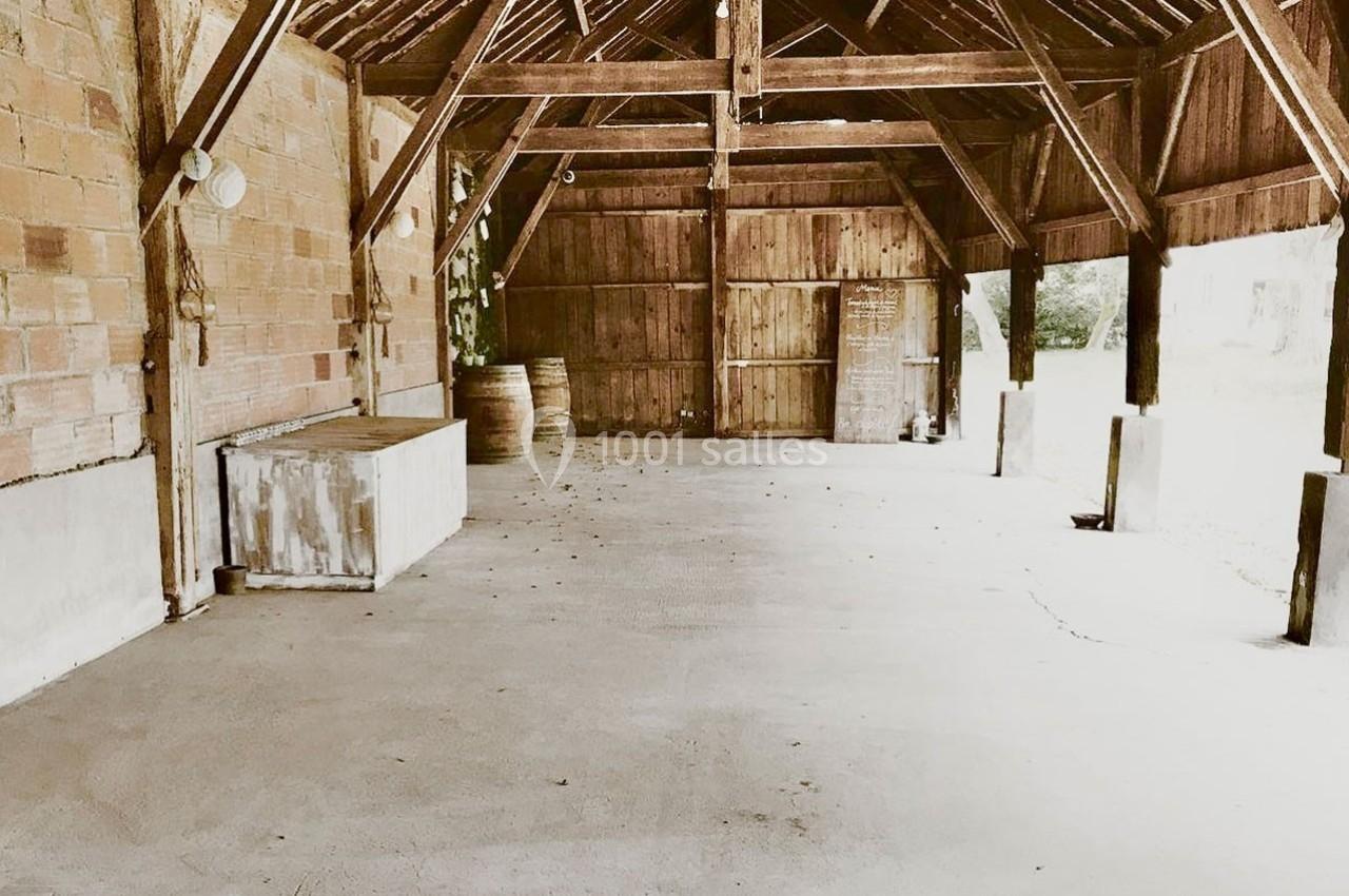 Abri en bois ouvert avec toit en poutres apparentes, sol en béton et quelques tonneaux en arrière-plan.