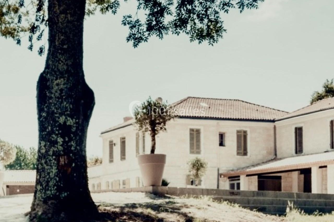 Bâtiment en pierre avec toit en tuiles, entouré d'arbres et d'un grand pot de fleurs dans une cour ensoleillée.