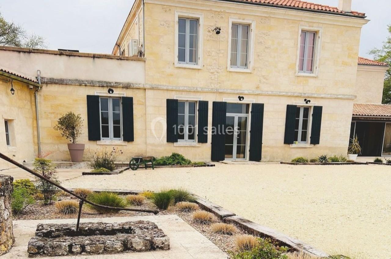 Façade d'une maison en pierre avec volets noirs, cour gravillonnée et aménagements paysagers simples au premier plan.