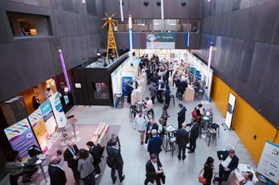 Miniature Agence Ilago #2 Des personnes échangent dans un espace d'exposition intérieur avec des stands et une structure métallique visible en arrière…