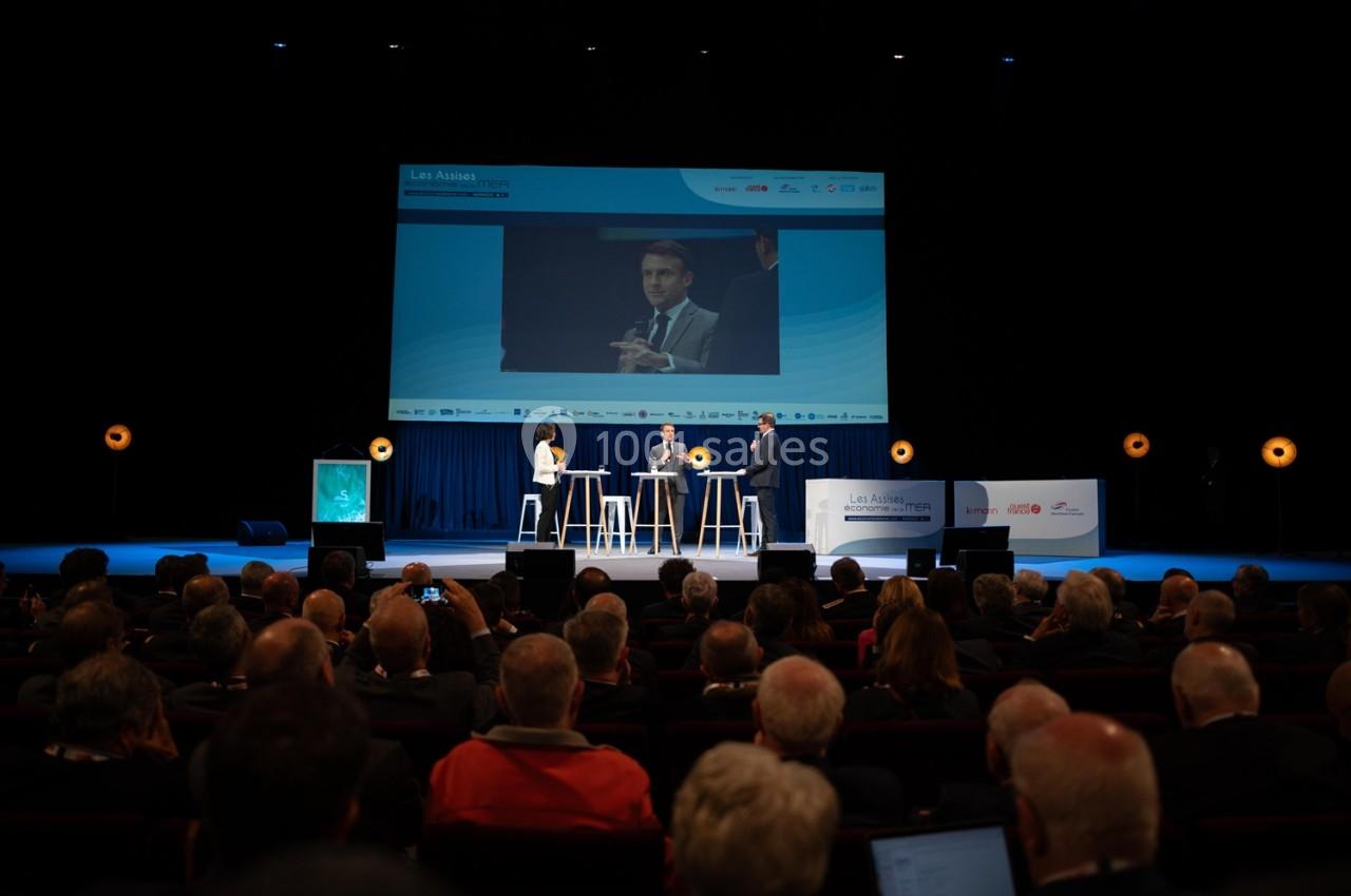 Conférence avec des intervenants sur scène, retransmise sur un écran géant devant un public attentif.
