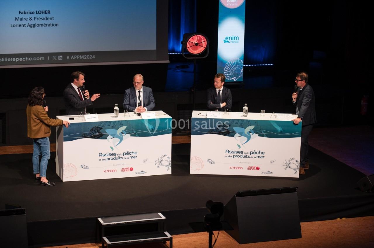 Cinq personnes discutent sur une scène lors d'une conférence, avec des tables et un écran en arrière-plan.