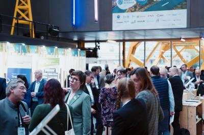 Miniature Agence Ilago #6 Des personnes échangent dans un espace d'exposition intérieur avec des stands et une structure métallique visible en arrière…