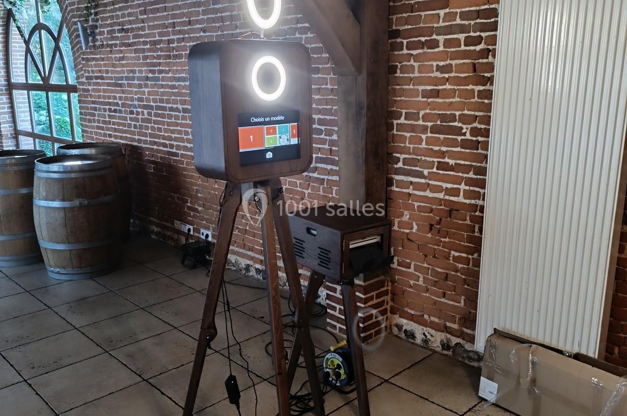 Borne photo sur trépied avec anneau lumineux, installée dans une pièce en briques avec tonneaux en arrière-plan.