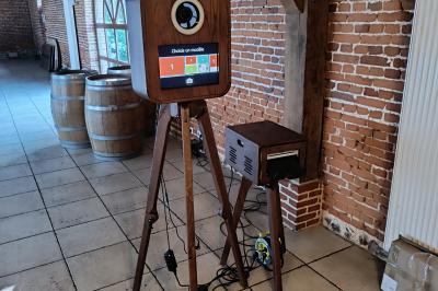 Borne photo sur trépied avec anneaux lumineux, installée dans une salle en briques avec tonneaux en arrière-plan.