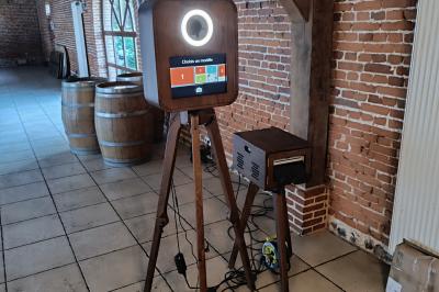 Borne photo sur trépied avec anneaux lumineux, installée dans une salle en briques avec tonneaux en arrière-plan.