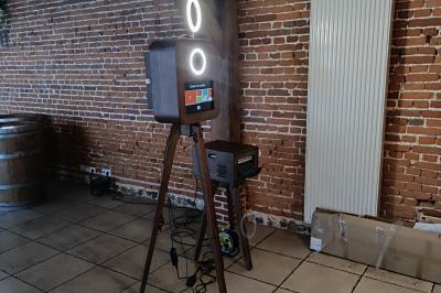 Borne photo sur trépied avec anneaux lumineux, installée dans une salle en briques avec tonneaux en arrière-plan.