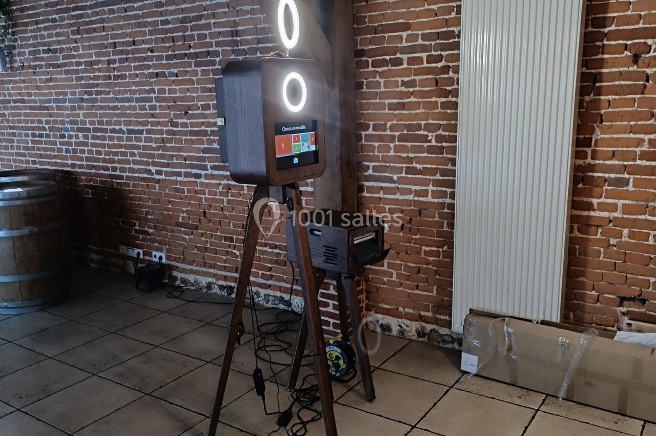 Borne photo sur trépied avec éclairage circulaire, installée dans une pièce aux murs en briques et sol carrelé.