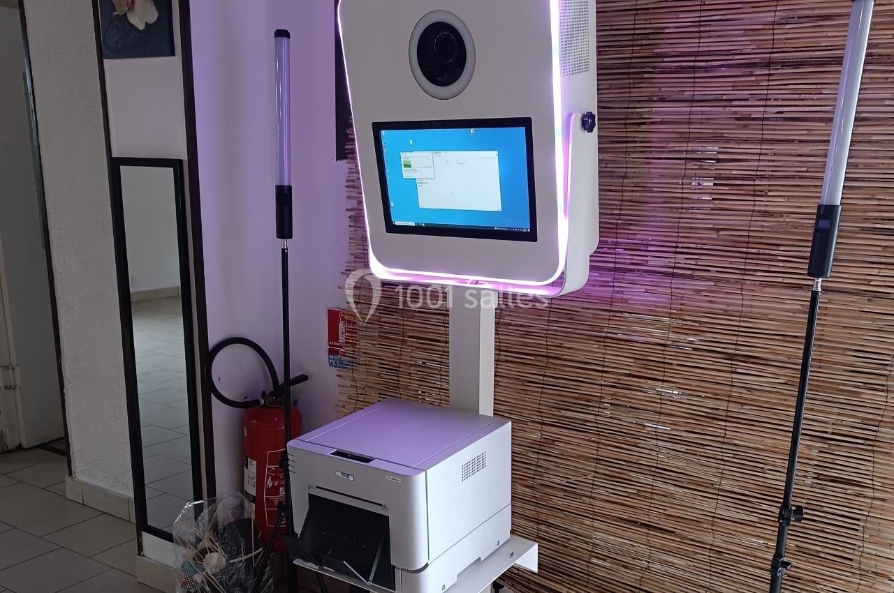 Borne photo avec écran tactile et imprimante intégrée, installée devant un mur en bambou dans un espace intérieur.