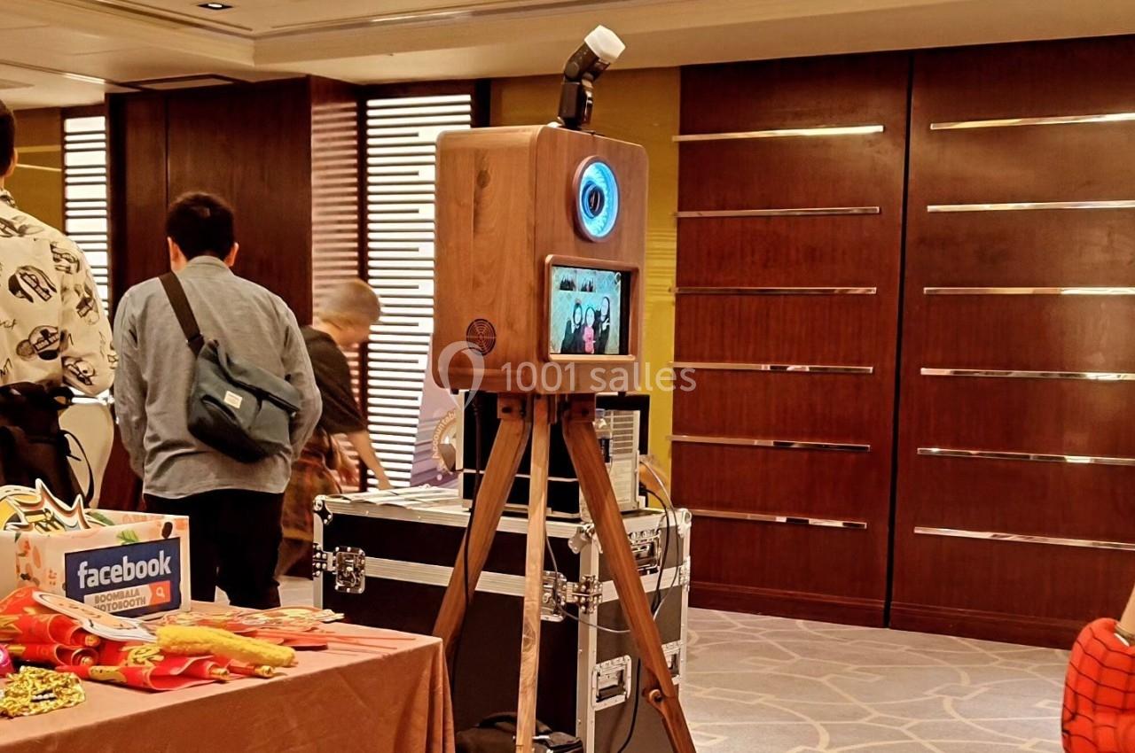 Cabine photo en bois sur trépied avec écran et flash, installée dans une salle avec des tables et des participants.