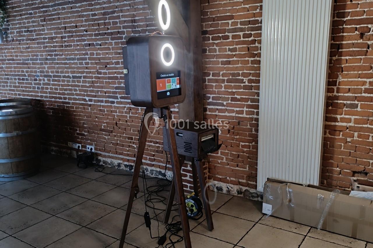Borne photo sur trépied en bois avec écran tactile et anneaux lumineux, installée dans une pièce aux murs en briques.