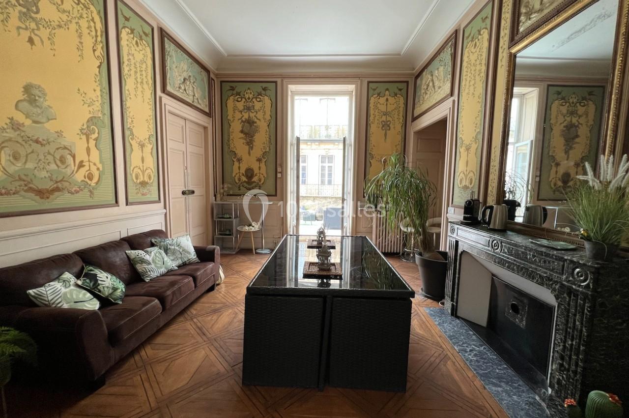 Salon élégant avec parquet en bois, canapé marron, table en verre, cheminée en marbre et murs ornés de fresques décoratives.