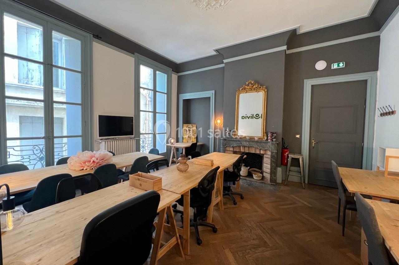 Salle lumineuse avec de grandes fenêtres, tables en bois, chaises noires et un miroir doré au-dessus d'une cheminée.