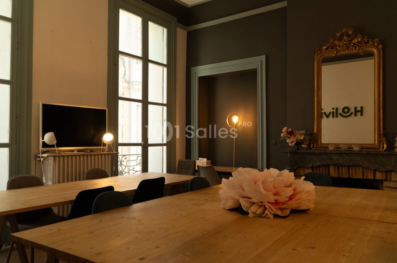 Salle lumineuse avec de grandes fenêtres, tables en bois, décoration sobre et une fleur en centre de table.