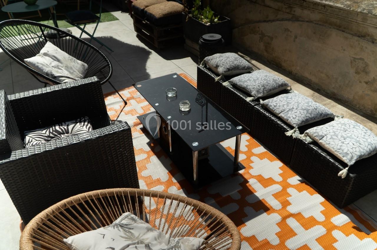 Mobilier de jardin avec fauteuils en rotin et coussins, table basse noire et tapis orange à motifs géométriques.