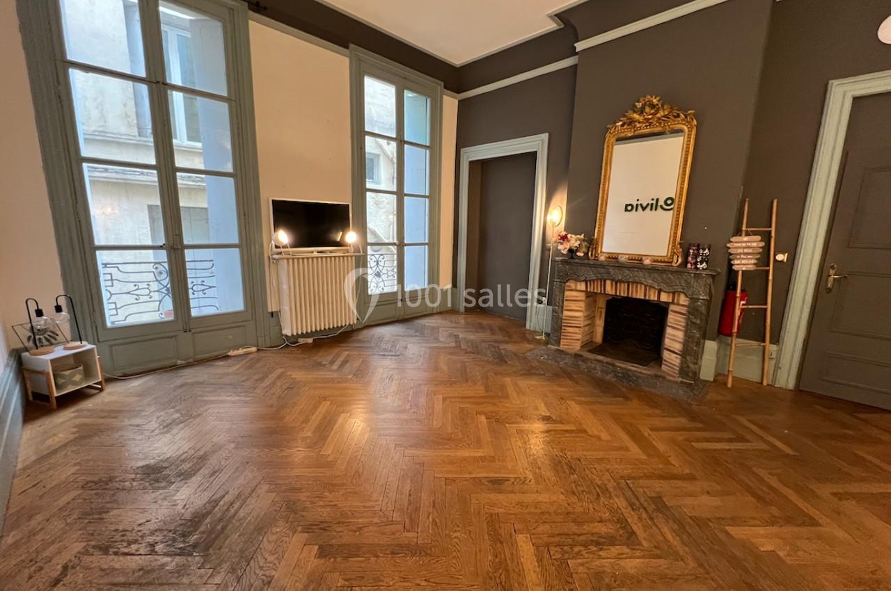 Salon lumineux avec parquet en chevrons, cheminée ornée d'un miroir doré et grandes fenêtres donnant sur l'extérieur.