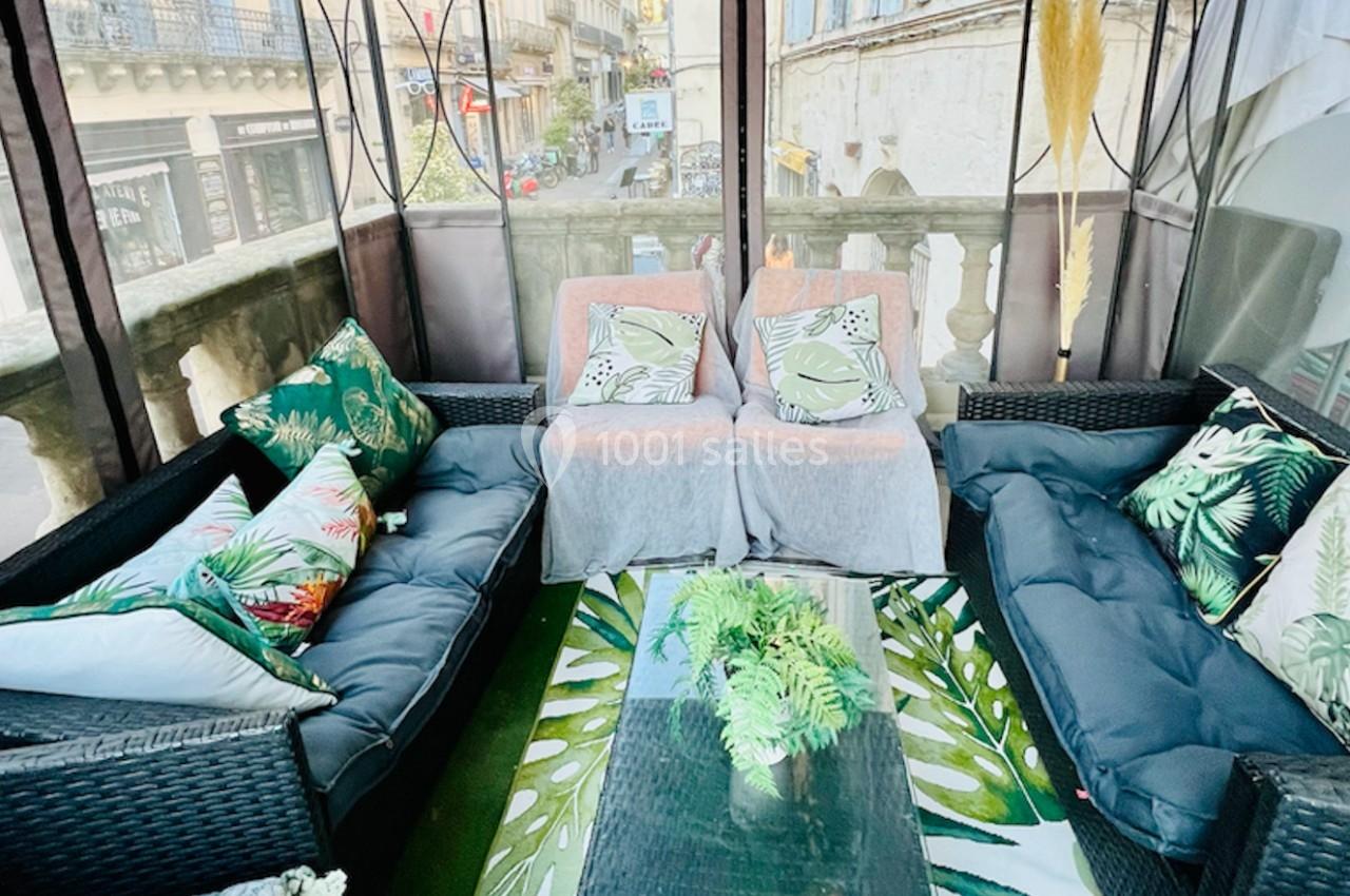 Salon extérieur couvert avec canapés en rotin, coussins à motifs tropicaux et vue sur une rue urbaine.