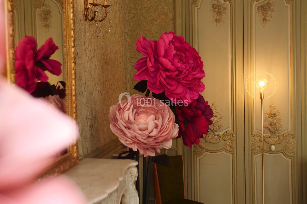 Grandes fleurs décoratives roses et rouges disposées dans une pièce élégante avec moulures et éclairage tamisé.