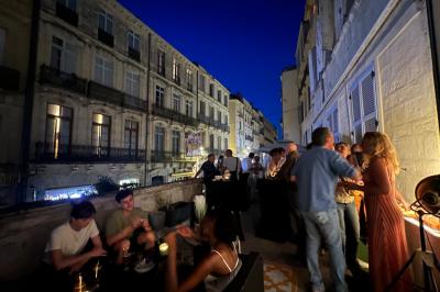 Groupe de personnes discutant et partageant un moment convivial sur une terrasse en soirée, avec des immeubles en arrière…