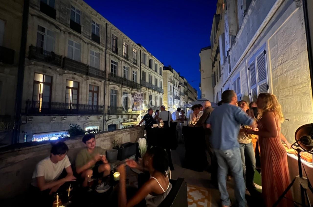 Groupe de personnes discutant et partageant un moment convivial sur une terrasse en soirée, avec des immeubles en arrière…