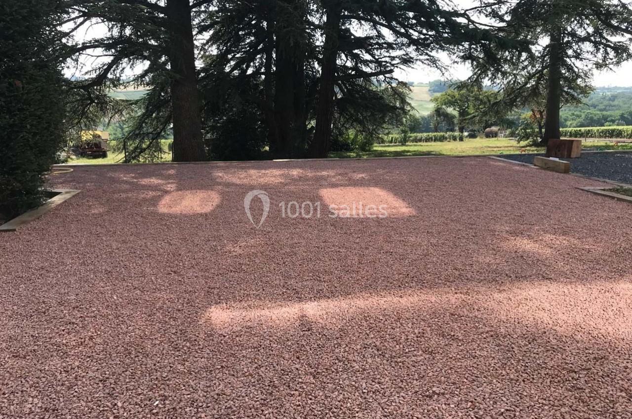 Allée en gravier rouge entourée d'arbres, avec vue sur un paysage rural en arrière-plan.