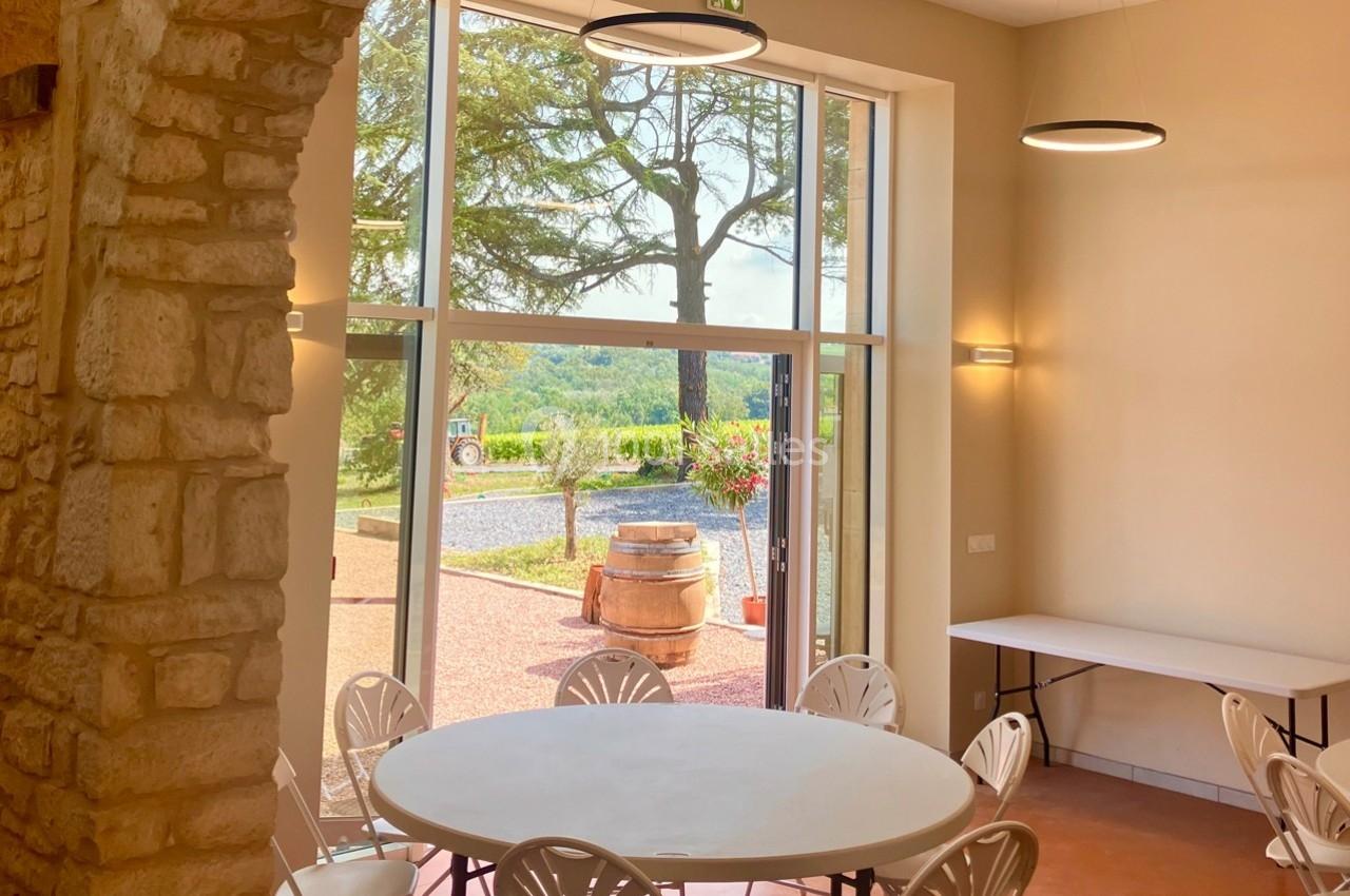 Salle lumineuse avec des tables rondes, vue sur une terrasse et un paysage verdoyant à travers de grandes baies vitrées.