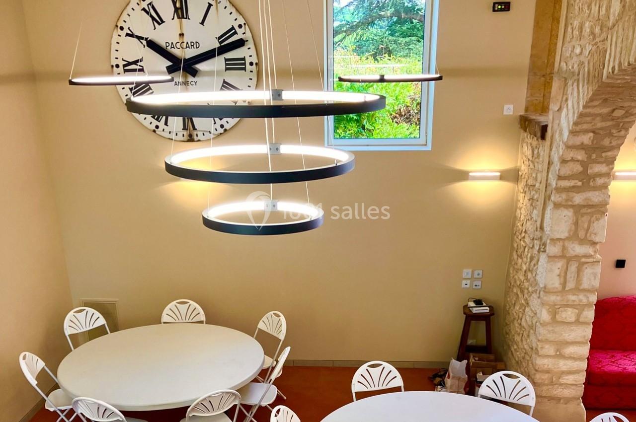 Salle lumineuse avec tables rondes blanches, chaises assorties, suspension moderne et grande horloge murale.