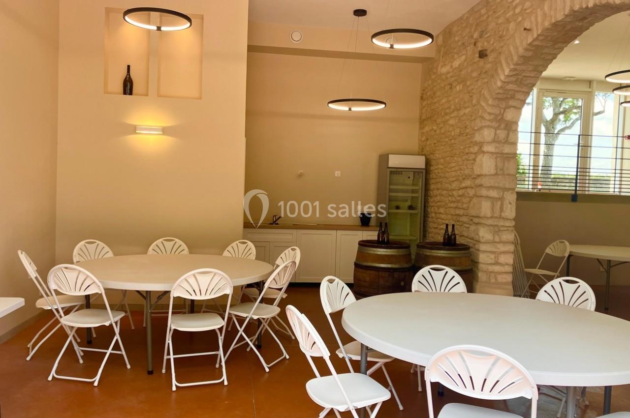 Salle lumineuse avec murs en pierre, tables rondes blanches, chaises pliantes et quelques bouteilles exposées.