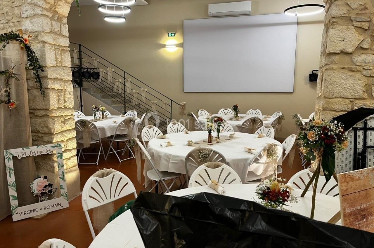 Salle décorée pour un événement, avec des tables rondes dressées, des chaises blanches et des arrangements floraux.