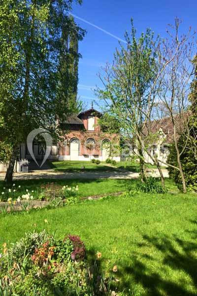 Jardin verdoyant avec pelouse, fleurs et arbres, devant une maison en briques sous un ciel bleu dégagé.